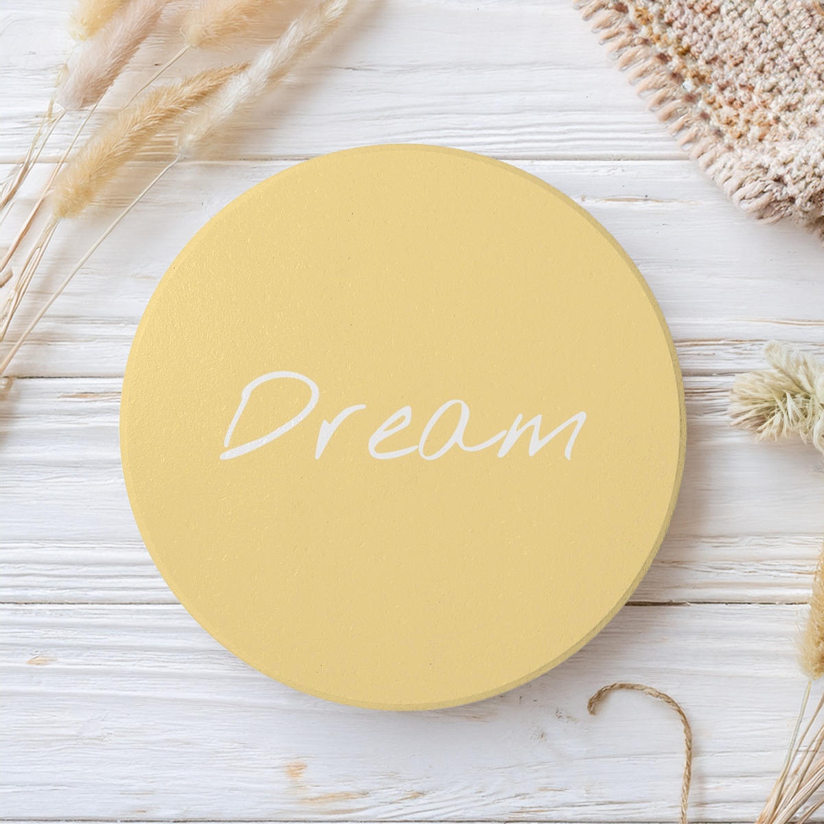 A round yellow coaster with the word 'Dream' written in white script, displayed on a white wooden surface with decorative dried grass and a knit fabric.